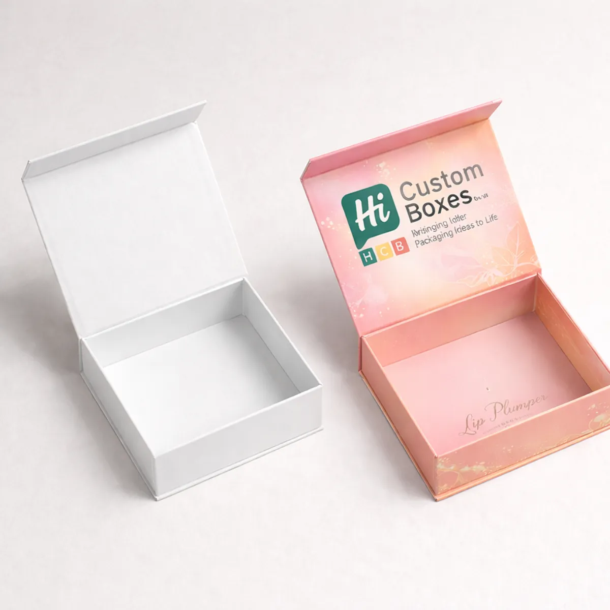 Two open square gift boxes with magnetic lids, featuring a white interior and pink branded design. class=